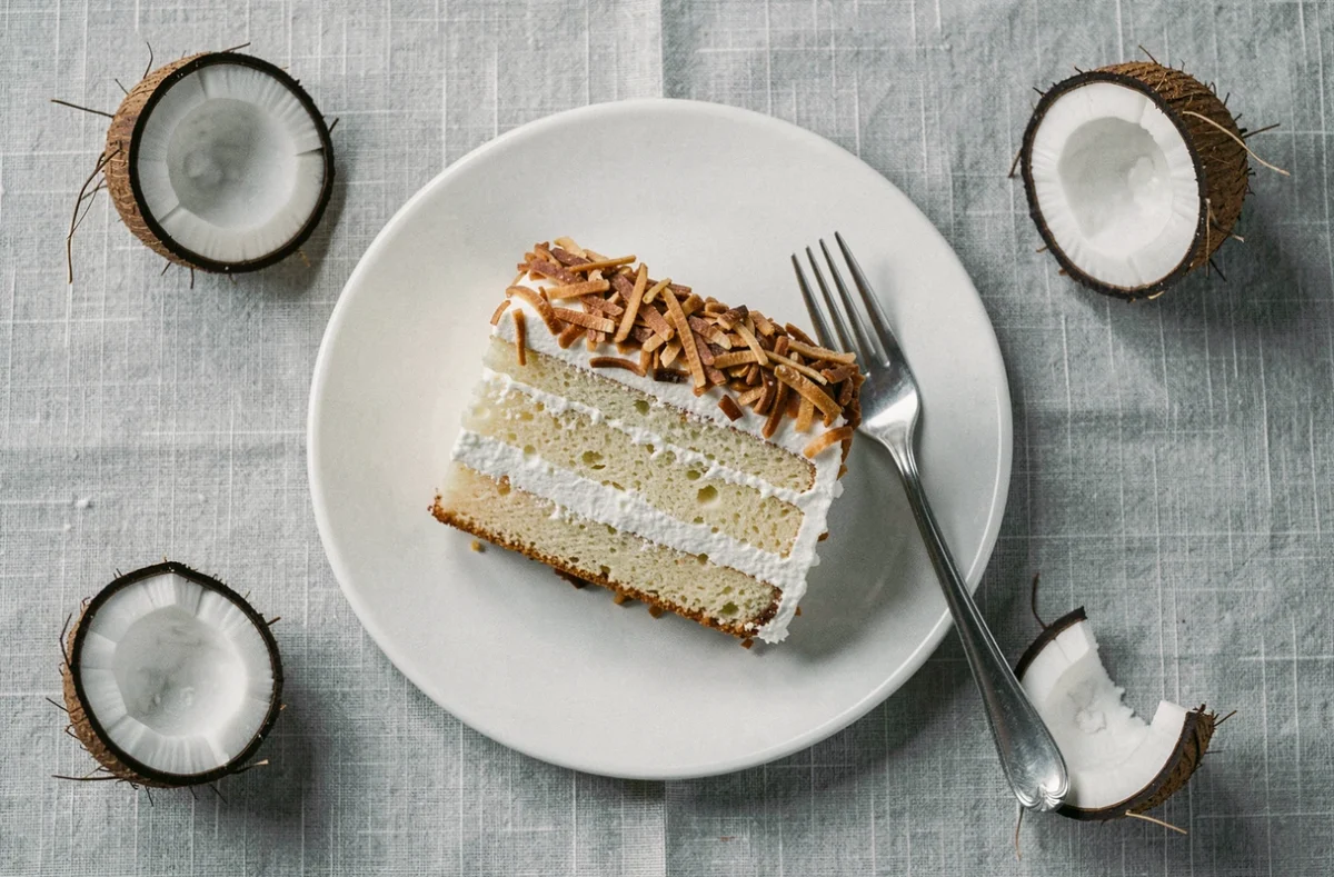 Tranche de gâteau à la noix de coco sur une assiette blanche vue de dessus avec décoration de noix de coco entière sur nappe grise